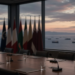 Empty diplomatic conference table at dusk overlooking distant tanker shipping lanes, conveying Middle East diplomacy and Hormuz risk without strike graphics, maps, or coordinates.