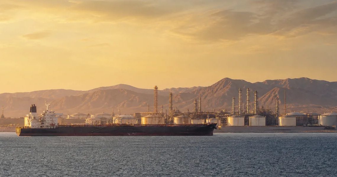 Oil tanker passing a coastal energy terminal under desert mountains at golden hour, illustrating southern Saudi energy and Red Sea shipping risk.