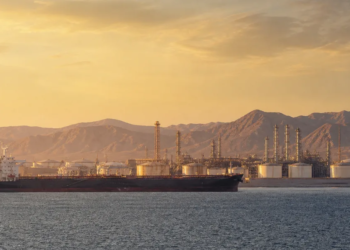 Oil tanker passing a coastal energy terminal under desert mountains at golden hour, illustrating southern Saudi energy and Red Sea shipping risk.