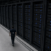 One unidentifiable technician, seen from behind, walking a hyperscale aisle beside a wall of GPU-style racks that dwarf him.