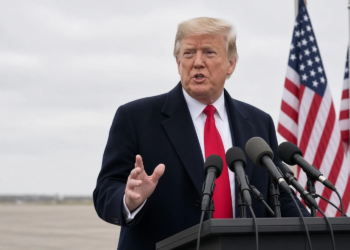 Trump speaking at a press podium, 3/4 front, mic cluster, United States flags behind.