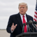 Trump speaking at a press podium, 3/4 front, mic cluster, United States flags behind.