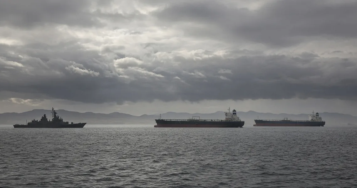 Warship and commercial oil tankers on a gray sea under heavy clouds, suggesting US-Iran tanker confrontation without graphic violence.