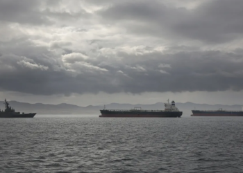 Warship and commercial oil tankers on a gray sea under heavy clouds, suggesting US-Iran tanker confrontation without graphic violence.