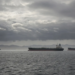 Warship and commercial oil tankers on a gray sea under heavy clouds, suggesting US-Iran tanker confrontation without graphic violence.