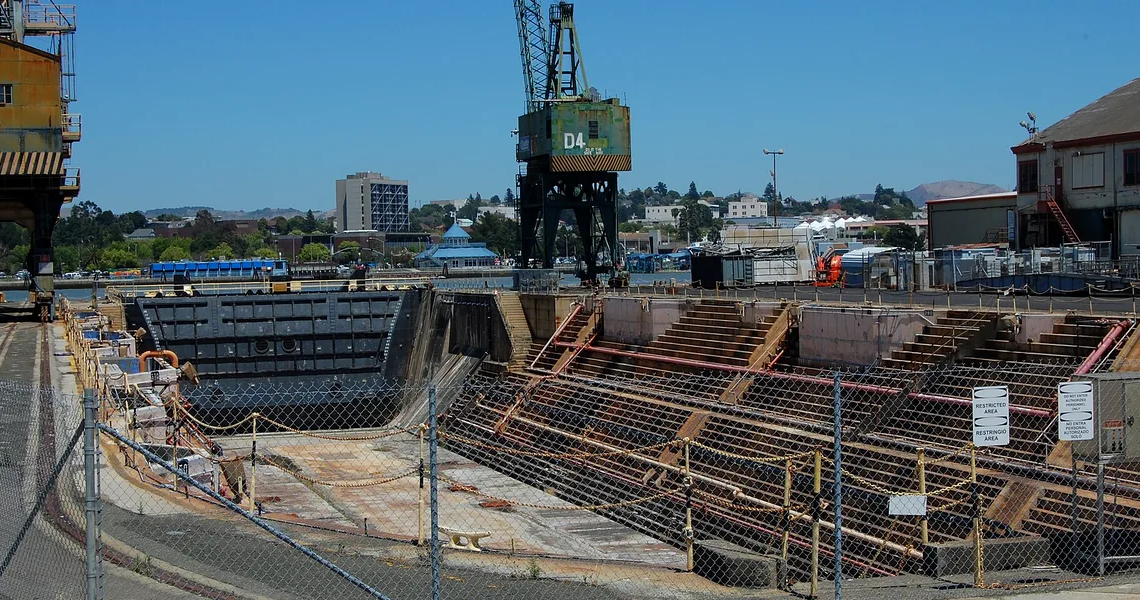 Naval shipyard dry dock with large steel hull and cranes, conveying shipbuilding acquisition competition.