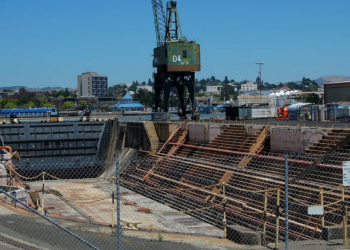 Naval shipyard dry dock with large steel hull and cranes, conveying shipbuilding acquisition competition.