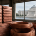 Stacked copper cathodes and coils in a warehouse with a distant neoclassical government building, conveying a stalled copper tariff plan without implying duties imposed.