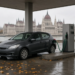 Compact hatchback parked PARALLEL to a pump island (side at the pump), green diesel nozzle in the side filler, Hungarian Parliament across the Danube.